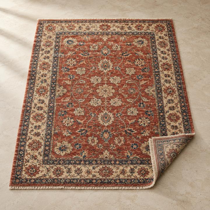 Rust-red rug with a dense floral pattern and patterned border, one corner turned up